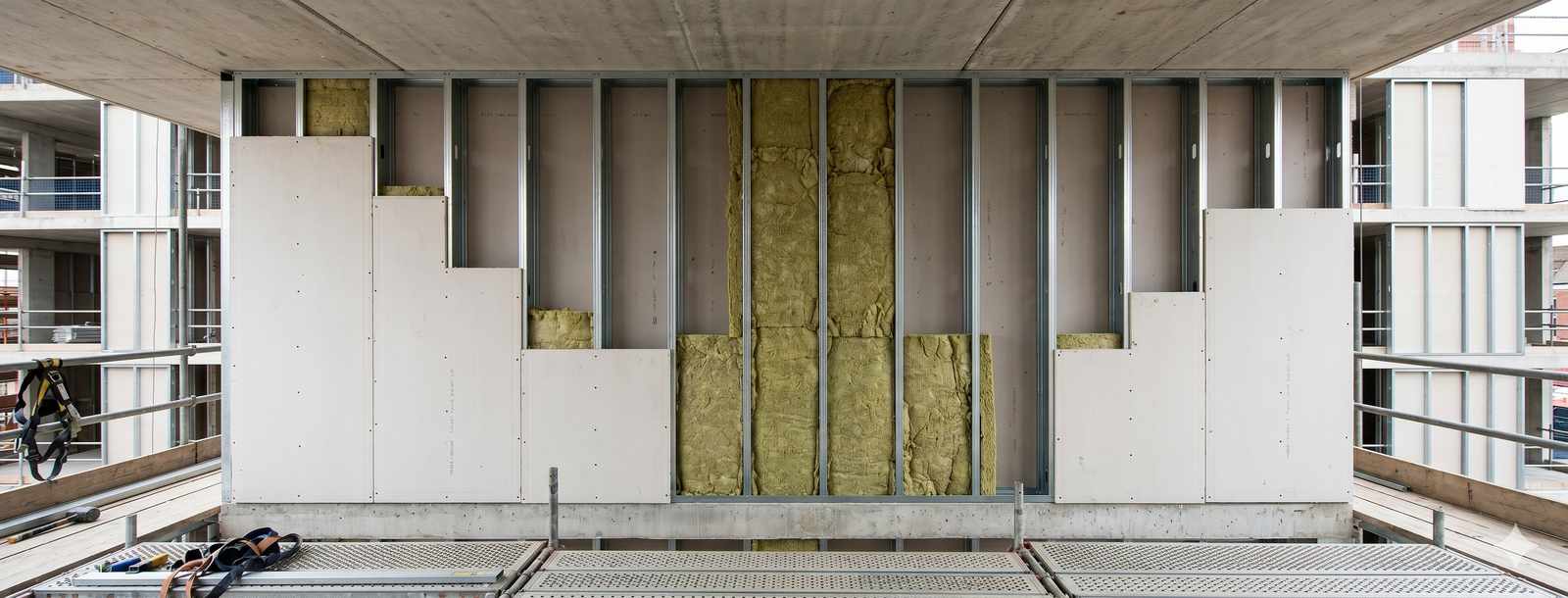 Close-up architectural photograph of cold-formed light gauge steel C-studs and U-tracks neatly stacked on a UK construction site, with the characteristic perforation pattern visible on the C-section webs. Bright daylight raking across the steel surfaces showing profile detail. Sharp focus on the stud sections, shallow depth of field with construction site context blurred in background. Documentary photography style. No supplier logos visible.